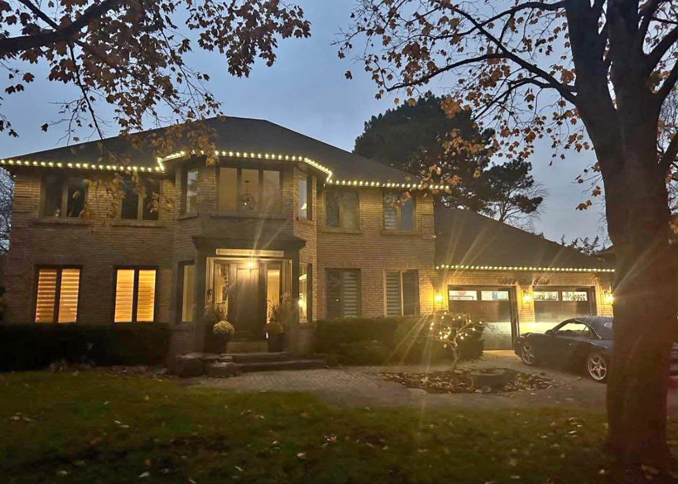 Large brick home at sunset with warm white lights along its long rooflines, framed by mature trees
