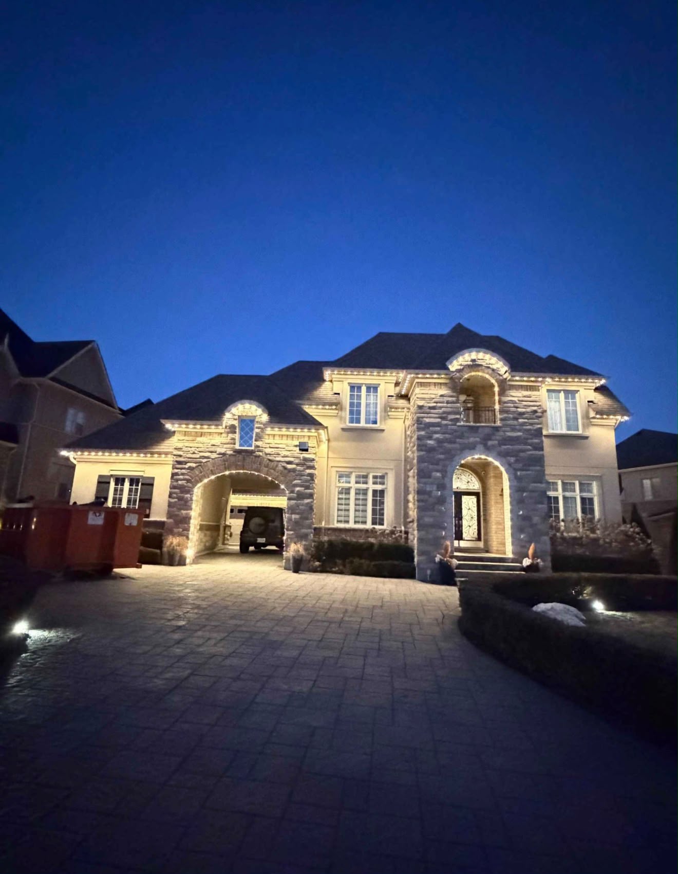 Large stone-front home at blue hour with warm white lights following its hipped rooflines and front entry arch