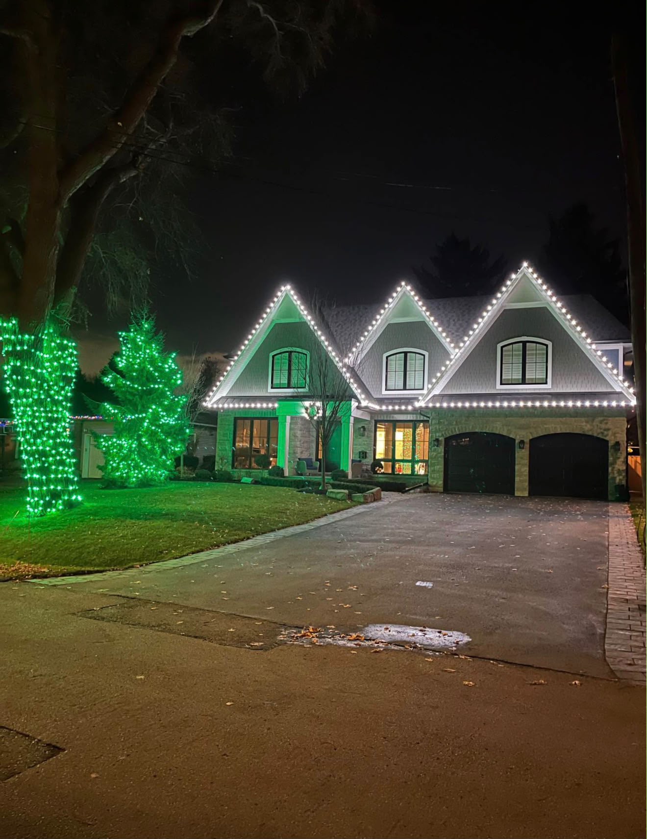 Home with three white-lit peaked gables and two front-yard trees wrapped in bright green lights